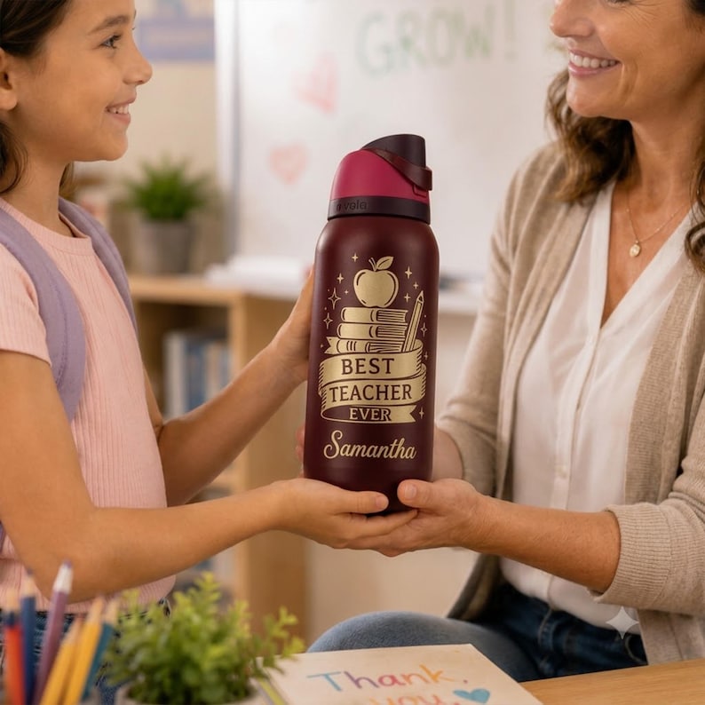 May include: A maroon water bottle with a gold design featuring an apple, books, and a pencil. The text reads "BEST TEACHER EVER" and "Samantha". The bottle has a dark red lid and is being held by two people.