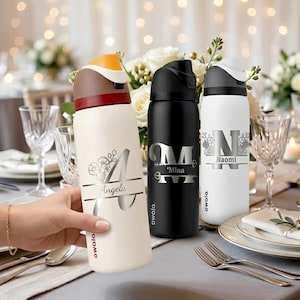 Puede incluir: Tres botellas de agua Owala con nombres personalizados y dise&ntilde;os florales o de flechas. Una es crema con tapa marr&oacute;n y amarilla, otra es negra con tapa negra y otra es blanca con tapa gris. Cada botella presenta una letra grande y un nombre.