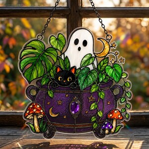 May include: A stained glass suncatcher featuring a purple cauldron filled with green monstera and pothos leaves, a white ghost, and a black cat. The cauldron is decorated with a purple gem, gold stars, and moons, with red and purple mushrooms below.