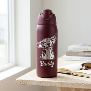 Puede incluir: Una botella de agua de acero inoxidable de color granate con tapa abatible. La botella presenta un gr&aacute;fico de un perro sonriente con el nombre "Buddy" impreso debajo. La botella est&aacute; sobre una mesa de piedra.