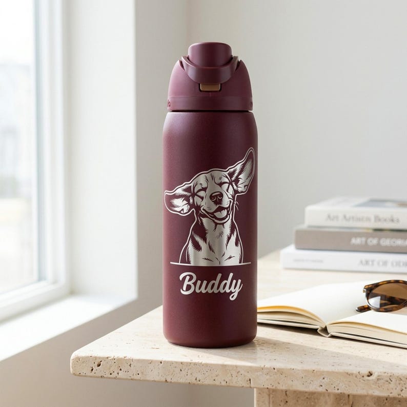 Puede incluir: Una botella de agua de acero inoxidable de color granate con tapa abatible. La botella presenta un gr&aacute;fico de un perro sonriente con el nombre "Buddy" impreso debajo. La botella est&aacute; sobre una mesa de piedra.