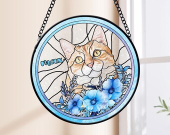 May include: A round stained glass window hanging with a chain. The window features a portrait of a cat with orange and white fur, surrounded by blue flowers. The name "Max" is written in the top left corner.