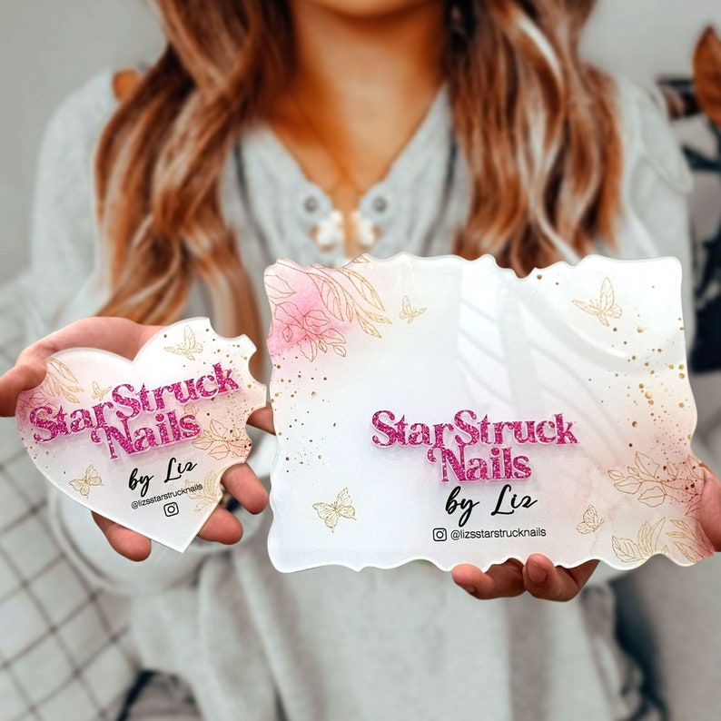 Op de afbeelding: Twee witte acryl borden met een roze en gouden bloemenontwerp. De borden tonen de tekst "StarStruck Nails by Liz" met een roze glitterafwerking. Het kleinere bord is hartvormig en het grotere bord is rechthoekig.