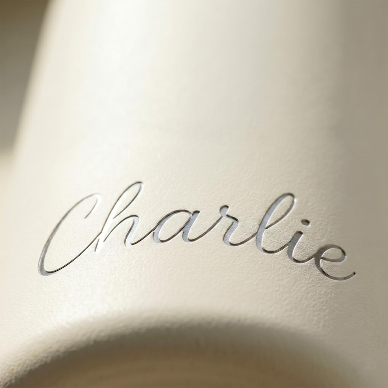 Puede incluir: Primer plano de una superficie color crema con el nombre "Charlie" grabado elegantemente en una fuente cursiva. La rotulaci&oacute;n es de un tono m&aacute;s claro, creando un contraste sutil.