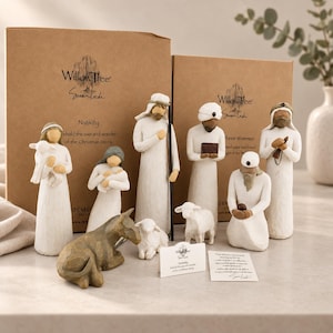 May include: A Willow Tree Nativity set featuring carved resin figurines of figures in white robes, including a shepherd with a lamb, a kneeling figure with a baby, a tall figure with a staff, three wise figures, a donkey, and a small lamb.