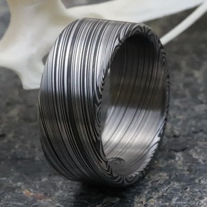 May include: A close-up of a wide, patterned ring. The ring features a unique design of vertical and curved lines in shades of gray and black. The ring's surface has a textured appearance, and the overall style is modern and artistic.