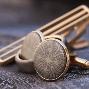 Könnte beinhalten: Ein Paar goldene Manschettenknöpfe mit einem gemusterten Design. Die Manschettenknöpfe sind rund und haben einen goldenen Rand. Das Muster ist ein wirbelndes, abstraktes Design in Grautönen.