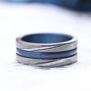 Könnte beinhalten: Ein silberner Ring mit einem blauen und silbernen Muster. Der Ring besteht aus drei übereinander gestapelten Bändern.