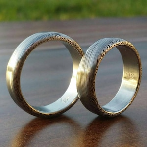 May include: Two silver wedding bands with a gold pattern. The bands are stacked on top of each other.
