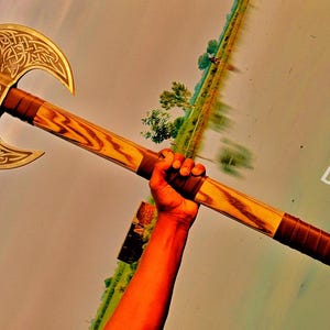 May include: A double-bitted battle axe featuring a gold-toned metal head with intricate Celtic knotwork engravings and a central skull emblem. The handle is crafted from polished wood with dark brown leather-wrapped grips and the text DamasteelheritageCo.