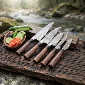 Pode incluir: Um conjunto de cinco facas de cozinha com lâminas de metal martelado e cabos de madeira escura, dispostas por ordem decrescente de tamanho. As facas estão sobre uma tábua de madeira envelhecida com o texto DamasteelheritageCo e uma tigela.