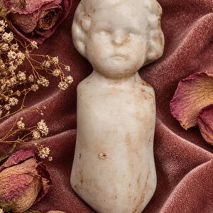 May include: A vintage porcelain doll torso featuring a sculpted head with molded hair and facial features. The off-white material displays visible signs of age, including brown staining and surface wear across the chest and face area.