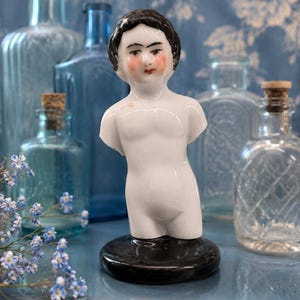 May include: A white porcelain doll torso featuring a painted face with black hair, arched eyebrows, and rosy cheeks. The figure is mounted on a circular black base, showcasing a smooth, glossy finish and a classic, upright, armless posture.