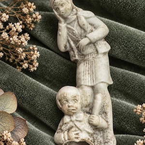 May include: A light-coloured, weathered figurine depicting two figures: one standing figure with a hand to their face and a second figure positioned below, appearing to look upward. The sculpture has a textured, matte finish with visible signs of age.
