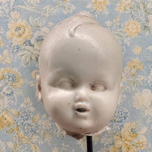 May include: A white porcelain doll head featuring molded hair with a single curl on the forehead, closed eyes, and a small open mouth. The surface shows minor dark spots and wear, and the head is mounted on a thin black metal rod for display.
