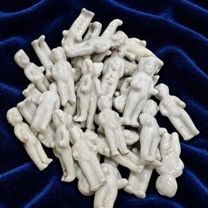 Könnte beinhalten: Eine Sammlung kleiner, weißer, unglasierter Porzellanfiguren in Form von menschlichen Puppen. Jede Figur weist eine einfache, geformte Gestalt mit definierten Gliedmaßen und einer glatten, glänzenden Oberfläche auf. Die Figuren liegen in einem Haufen.