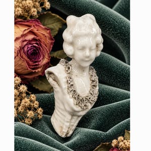 May include: A white ceramic bust figurine depicting a person with a detailed hairstyle and a textured, ruffled collar. The sculpture features a smooth, glossy finish and is sculpted from the chest up, showcasing intricate facial features.