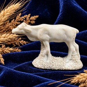 May include: A white, carved figurine of a standing cow or calf, positioned on a textured, rocky-looking base. The animal is depicted in profile, facing left, with a smooth, matte finish and visible muscular definition along its body.