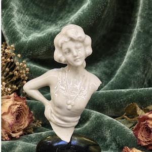 May include: A white ceramic bust figurine depicting a person with short wavy hair, wearing a necklace with three circular pendants. The figure is posed with one hand resting on the hip and is mounted on a smooth, rounded black base.