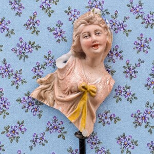 May include: A porcelain half-doll figurine featuring a person with light brown hair, a peach-colored bodice, and a bright yellow bow at the center. The figure has painted facial features, including blue eyes and red lips, and is mounted on a thin black rod.