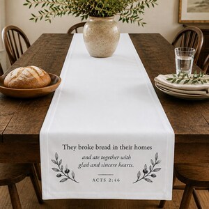 May include: A white fabric table runner featuring the black printed text, "They broke bread in their homes and ate together with glad and sincere hearts. ACTS 2:46," flanked by two black botanical illustrations of leafy branches on the ends.