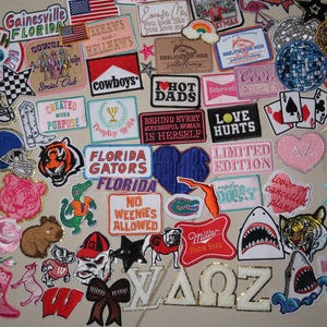 May include: A large collection of assorted embroidered fabric patches featuring various designs including sports team logos, text phrases, animals, hearts, stars, and collegiate Greek letters in a wide array of vibrant colors and shapes.