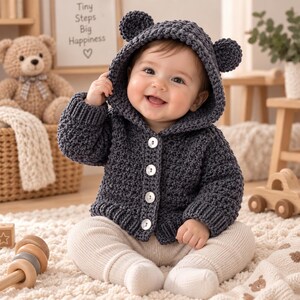 Peut inclure: Un gilet à capuche en crochet gris anthracite foncé doté de deux petites oreilles d'ours arrondies sur la capuche et de quatre boutons blancs sur le devant. Le vêtement présente un motif texturé, des manches longues et des bords côtelés.