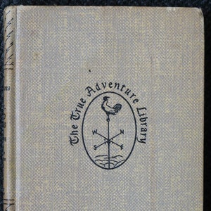 May include: A tan book with a black and white logo on the cover. The logo is a circle with a rooster on top of a compass. The text around the logo reads "The True Adventure Library".