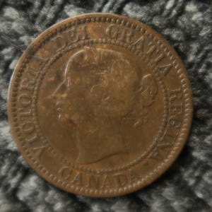 May include: A close-up of a copper coin featuring a woman's profile. The coin is inscribed with "VICTORIA DEI GRATIA REGINA CANADA". The coin is set against a textured grey background.