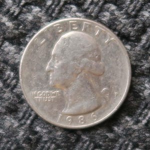 Peut inclure: Une pièce de monnaie américaine en argent d'un quart de dollar avec l'année 1986 inscrite en bas. La pièce représente un profil de George Washington et les mots "Liberty" et "In God We Trust".