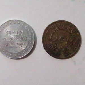 May include: Four different silver and bronze colored tokens. The first token is silver and features a pyramid with the text "Pennzoil  Super  Car  Oil". The second token is silver and features the text "Shell's  Mr. President  Coin Game". The third token is bronze and features the text "Dayland  D&J  1976  Coin Game". The fourth token is bronze and features a design of a person's head.