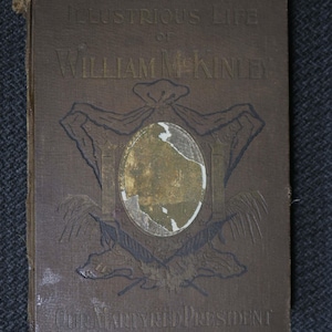 May include: A vintage book titled "Illustrious Life of William McKinley" with embossed text and a central oval design. The cover is a muted brown with gold accents and the words "Our Martyred President".