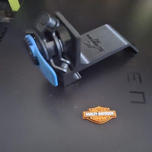 以下が含まれることがあります： 3Dプリントされた黒いL字型ブラケット。青いレバー機構と、オレンジと黒のハーレーダビッドソン・モーター・カンパニーの盾型エンブレムが付いています。ブラケットの表面には浮き彫りの文字があり、エンブレムにはブランド名がはっきりと表示されています。