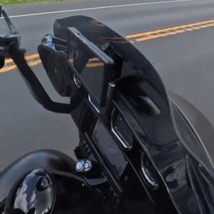 Può includere: Una carenatura per moto nera dotata di parabrezza oscurato, schermo digitale centrale e supporto per smartphone. Il gruppo include altoparlanti circolari integrati e un serbatoio del carburante nero visibile in primo piano.