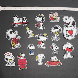 May include: A collection of fifteen embroidered Snoopy and Peanuts character patches arranged on a dark surface, with a measuring tape at the top showing a length of 15 inches. The patches feature various scenes including Snoopy in a red car, on a skateboard, wearing sunglasses, and holding a red heart, with one patch reading I Love Coffee.