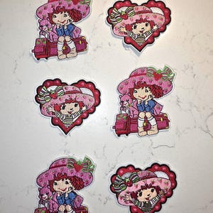 May include: Six embroidered patches featuring a cartoon character with red hair wearing a pink strawberry-patterned hat. Three patches show the character sitting on a pink suitcase with a small pink animal, and three are heart-shaped portraits.