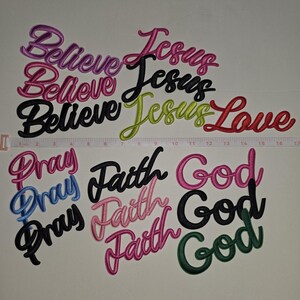 May include: A collection of embroidered word patches including Believe, Jesus, Love, Pray, Faith, and God in various colors like pink, black, lime green, red, blue, and dark green. A white measuring tape shows the patches span up to 17 inches wide.