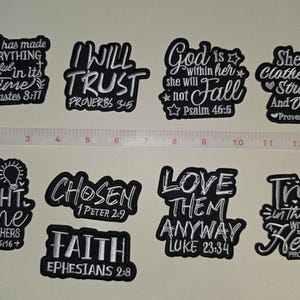 May include: A collection of nine black and white embroidered patches featuring religious text and scripture references, arranged in two rows next to a white measuring tape that shows a width of approximately 14 inches across the entire display.