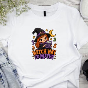 May include: A white t-shirt featuring a graphic of a cartoon witch with orange hair wearing a black hat, holding a purple calculator. The design includes a yellow crescent moon, stars, the numbers 1, 2, 3, 5, and the text Witch Way To Math?