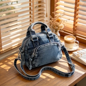 May include: A medium-sized denim handbag featuring a washed blue finish, two top handles, and a detachable shoulder strap. The bag includes a front zippered pocket with knotted pull tabs and a main zippered compartment with silver-toned hardware.