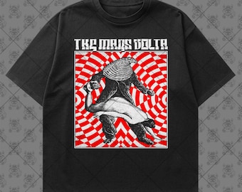 May include: A black short-sleeve t-shirt featuring a graphic print with the text THE MARS VOLTA. The design shows a figure with a large seashell for a head interacting with a reclining human form against a red and white circular pattern.