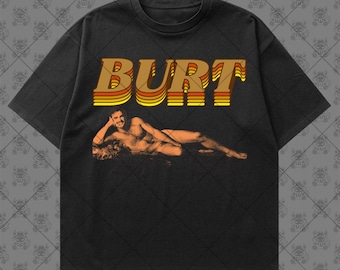 May include: A black short-sleeve t-shirt featuring the text BURT in a retro orange, yellow, and brown layered font. Below the text is a sepia-toned graphic of a person reclining on a surface, resting their head on one hand.