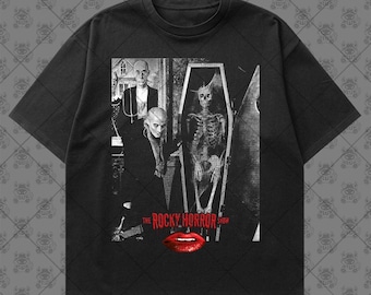 May include: A black short-sleeve t-shirt featuring a graphic print of a scene from The Rocky Horror Show. The print includes a black and white image of characters and a skeleton in a coffin, with the red text THE ROCKY HORROR SHOW and red lips.