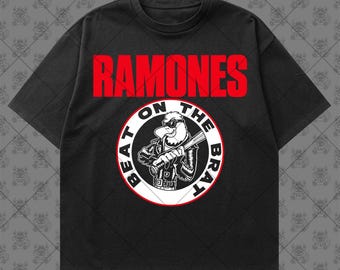 May include: A black short-sleeve t-shirt featuring the red text RAMONES above a circular graphic. The graphic depicts a white eagle wearing sunglasses and a leather jacket, holding a baseball bat, with the text BEAT ON THE BRAT.