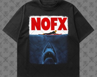May include: A black short-sleeve t-shirt featuring a graphic print of a person on a red surfboard above a large shark in deep blue water. The text NOFX is printed in bold red letters across the top of the graphic, set against a white background.