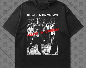 May include: A black short-sleeve t-shirt featuring a white graphic print of a crowd scene with the text DEAD KENNEDYS at the top and the text HOLIDAY IN CAMBODIA in red letters across the center of the graphic, set against a dark background.