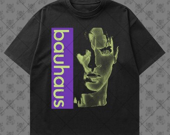 May include: A black short-sleeve t-shirt featuring a vertical purple rectangular block with the word "bauhaus" printed in light green. To the right of the text is a halftone graphic of a human face in light green, set against the black fabric.