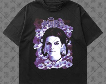 May include: A black short-sleeve t-shirt featuring a graphic print of a person surrounded by purple flowers. Above the person, the text reads "the MUFFS" in a stylized font. The design is centered on the chest of the garment.