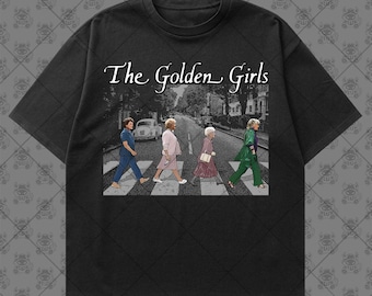May include: A black short-sleeve t-shirt featuring a graphic print of four figures walking across a crosswalk with the text The Golden Girls above them. The print depicts the characters in various outfits including blue, pink, and green clothing.