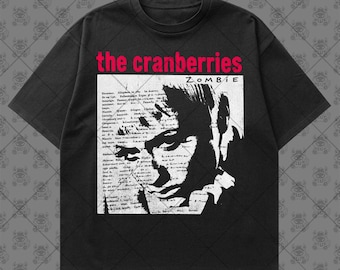 May include: A black short-sleeve t-shirt featuring a graphic print of The Cranberries band name in red text above a black and white image of a face overlaid with handwritten-style text and the word ZOMBIE in the upper right corner.